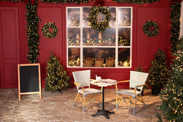 New Year's decorations of the street. Christmas trees with lights garlands, store window. Red decorated coffee house for christmas.  Xmas cafe terrace with table and chair. Cozy outdoor cafe on Xmas