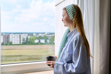 Young calm dreaming woman with cup in her hands, home window background