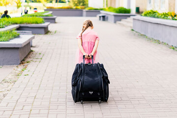 Full length shot of a little girl walking and pulling a suitcase. Back view of a little girl in a pink dress traveling with a big bag. A child walks and rolls a suitcase on wheels along the sidewalk.