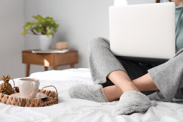 Fototapeta na wymiar Young woman in soft slippers resting on bed with modern laptop and cup of tea