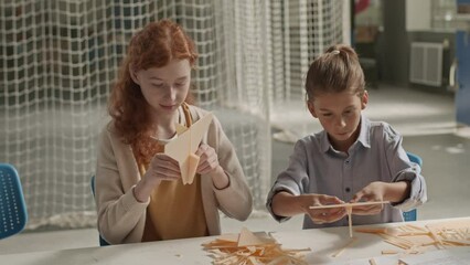 Waist up of two inquisitive preteen school classmates chatting while crafting wood airplane models at desk in technical club classroom