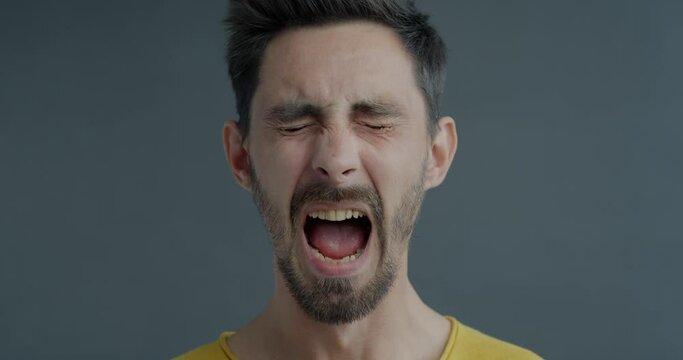 Slow motion close-up of young man screaming with anger and looking at camera on grey color background. People and negative emotion conceept.
