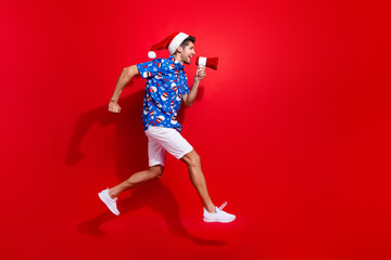 Fototapeta na wymiar Full size photo of funky young man jump run announcing toa wear santa claus print x-mas clothes isolated on red color background