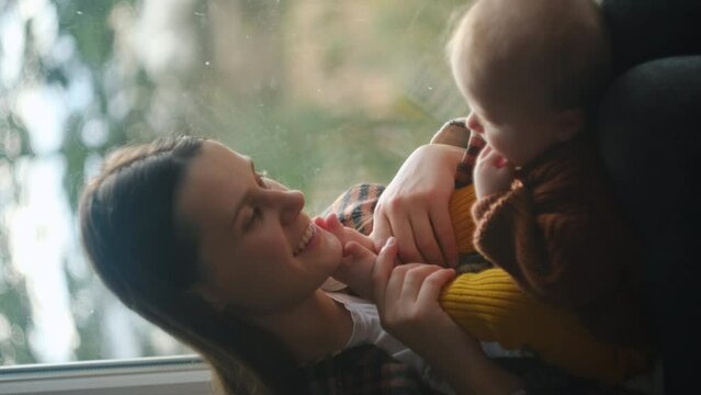 Vertical video of loving young mum with baby sitting on cozy windowsill at autumn nature background, talk to cute infant with warmth kiss foots. Affectionate mom caressing pampering little babe kid