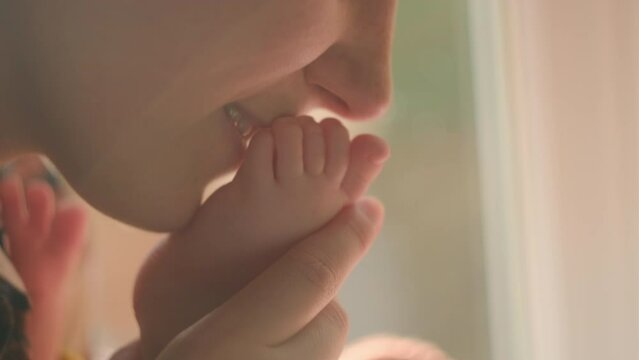 Close up of cheerful young mom gently kiss little feet in warm sunlight sitting on sill at home, tiny toes of newborn baby child. Tender moments with kid, mother and offspring connection, care concept