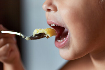 Close-up: a child's mouth with a spoon of mashed potatoes held up to it. Three-quarter turn.