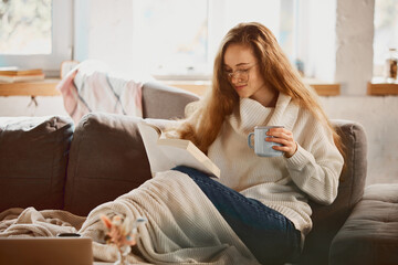 Young girl sitting on sofa, reading book and drinking hot fragrant warming tea after walking outside. Comfort at home. concept of lifestyle, winter holiday season, autumn weekend, relax atmosphere, - obrazy, fototapety, plakaty