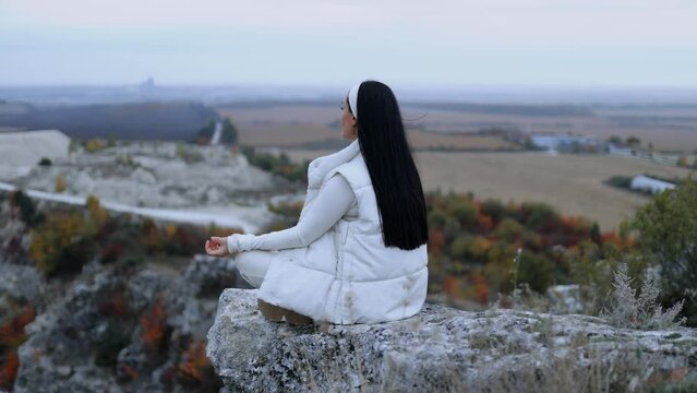 A young woman in a seated yoga position on the edge of a cliff, on top of a mountain, relaxing, looking and admiring the view. Woman meditating relaxation while traveling in autumn nature