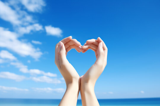 A woman's hands are stretched out in a heart shape against the blue sky. A concept suitable for celebrating happiness and being grateful for happiness, as well as a heart of kindness and warmth.