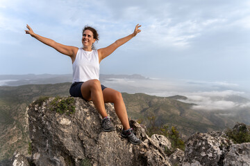 A young brunette caucasian woman with her arms open in joy sitting on a rock on the top of a...
