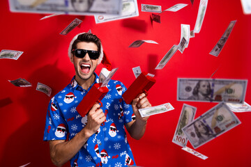 Photo portrait of funky young man shoot money gun luxury life dressed santa claus print x-mas clothes isolated on red color background