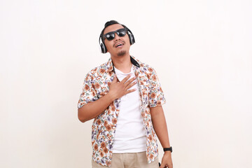 Relieved Asian man traveler standing while listening music using headphone. Isolated on white background with copyspace