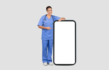 Happy european millennial doctor nurse in blue uniform, point finger at big phone with empty screen