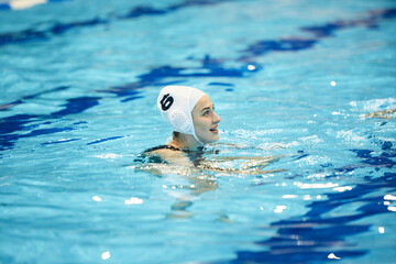 Happy young woman in cap swimming in pool with pure blue water and looking forwards during water...