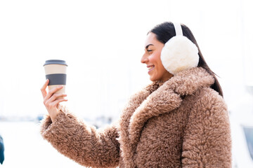Young woman wearing winter muffs  and holding take away coffee at outdoors with happy expression