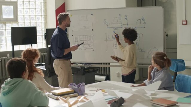 Medium shot of African American preteen schoolboy standing by whiteboard and pointing at aircraft construction schemes on it while answering to teacher at technical club