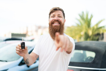 Redhead man with beard holding car keys at outdoors points finger at you with a confident expression