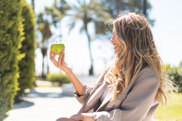 Pretty blonde Uruguayan woman with an apple at outdoors with happy expression