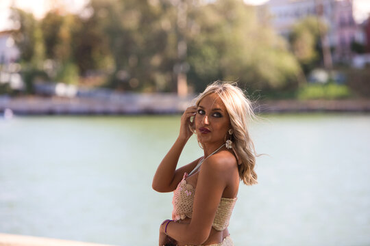 Young beautiful blonde woman dressed in embroidered trousers and crochet top by the river bank in seville. In the background a mediterranean style neighbourhood with white painted houses.