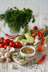 spices are ground in a small plate on a wooden board. vegetables around: zucchini, tomatoes, champignons, carrots, chili peppers, and greens in a glass, close-up, light background