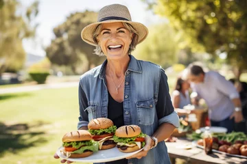 Poster Portrait of smiling mature woman eating hamburger at picnic in park © AI_images
