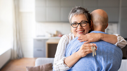 Happy senior woman hugging husband and holding home keys in hand - obrazy, fototapety, plakaty