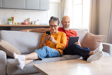 Happy Senior Spouses Using Modern Gadgets While Relaxing On Couch At Home