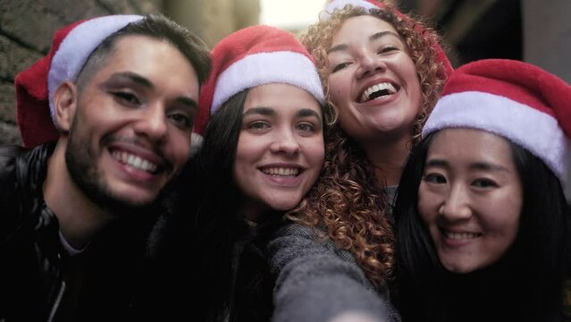 Diverse group of friends having fun celebrating Christmas on city street - Young people taking selfie video call wearing Santa Claus hats during holidays