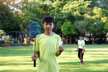 An Asian boy holds a badminton racket and a white shuttlecock while playing badminton with friends on the park lawn in the evening after returning from school.