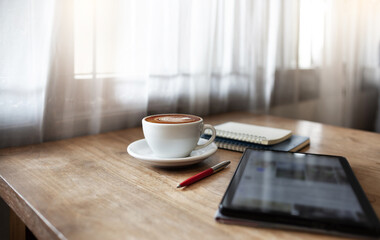 Latte coffee with tablet computer and note book on wooden table by bright window, people relaxing by using mobile phone for entertainment and social media online at cafe after work