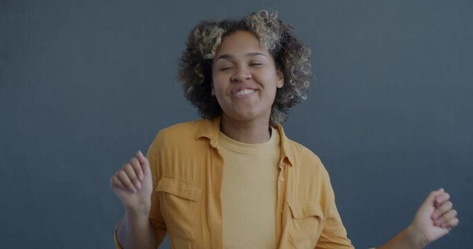 Slow motion portrait of joyful African American woman dancing listening to music looking at camera on gray background. People and youth culture concept.