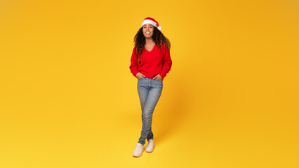 Joyful young black lady in Santa hat posing on yellow background, full length