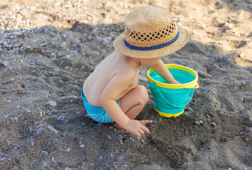 cute adorable baby boy kid is playing on beach with bucket and toy watering can.child with sun hat cat on head on seashore.sea waves summer vacation time.sunshine of face,warm weather