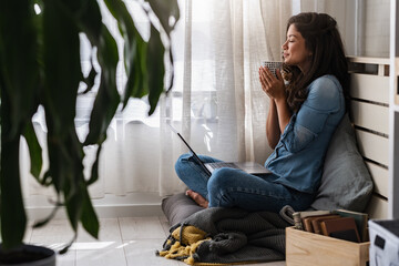 Beautiful smiling young woman sitting at home in a cozy window corner, drinking coffee, relaxing, enjoying a morning moment of peace