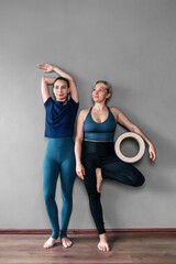 Overjoyed full-length female yoga trainers in sportswear stand barefoot near the wall, one holds a wooden circle in her hand, rejoicing at the start of classes or relaxing after the end of the lesson