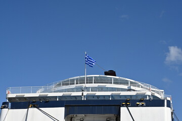 Aft part of passenger ferry with blue and white hull and Greece flag on top is observed against clear sky. Vessel is moored in port of Heraklion in Crete.