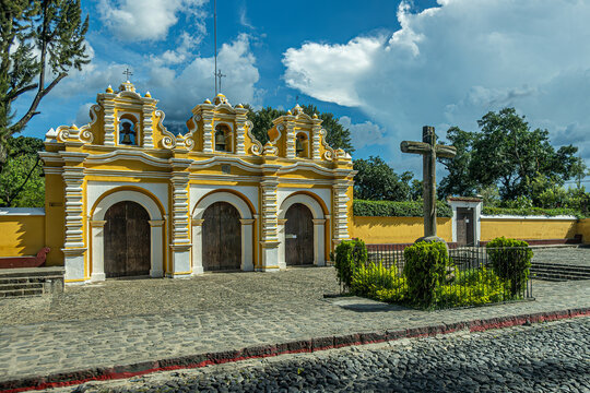 Guatemala, La Antigua - July 20, 2023: Iglesia El Calvario, Calvary Church, white and yellow monumental entrance to park in front with giant cross adjacent to street. Blue cloudscape and foliage