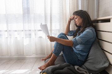 Stressed young woman holding financial papers sitting on the floor concerned about her finances - obrazy, fototapety, plakaty
