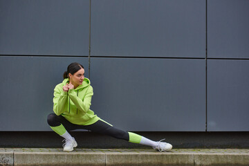 young sportswoman in hoodie and leggings doing lunges near grey wall outdoors, energy and sport - obrazy, fototapety, plakaty