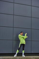 pretty sportswoman in hoodie and leggings warming up near grey wall outdoors, fresh air and sport