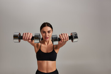 young motivated sportswoman in black active wear working out with dumbbells on grey background