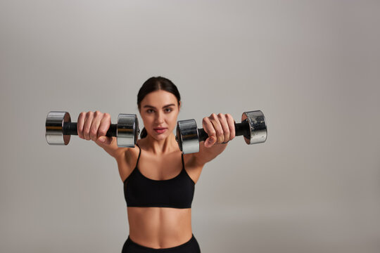 young motivated sportswoman in black active wear working out with dumbbells on grey background © LIGHTFIELD STUDIOS