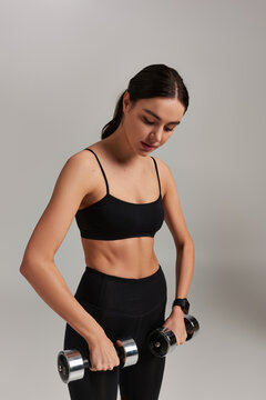 motivated and strong sportswoman in black active wear exercising with dumbbells on grey backdrop