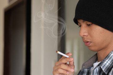 Asian boy sitting in front of dark wall of the toilet and smoking seriously, soft and selective focus, homeless and drug addiction concept.