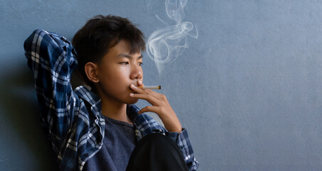 Asian boy sitting in front of dark wall of the toilet and smoking seriously, soft and selective focus, homeless and drug addiction concept.