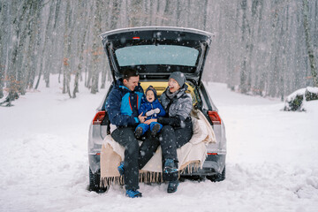 Laughing mom, dad and little girl are sitting on a blanket in the trunk of a car under a snowfall...