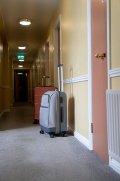 Rolling suitcase in a hotel hallway painted in pastel colors with gray carpeting in an older building in Dublin, Ireland. Hallway lighting switched on and daylight through an invisible window.