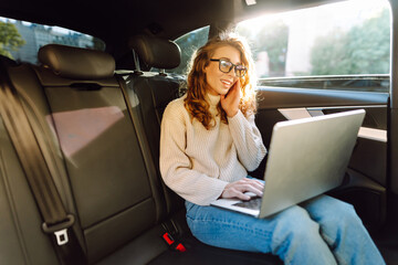 Young woman with a laptop in the back seat of a car. Business woman working on a laptop in a car. Freelance, business concept.