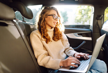 Young woman with a laptop in the back seat of a car. Business woman working on a laptop in a car. Freelance, business concept.