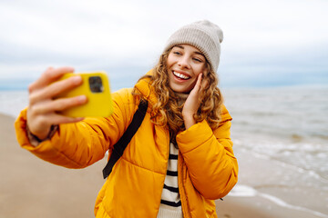 Happy woman in a bright jacket on the seashore with a phone in her hands. Young tourist woman takes...
